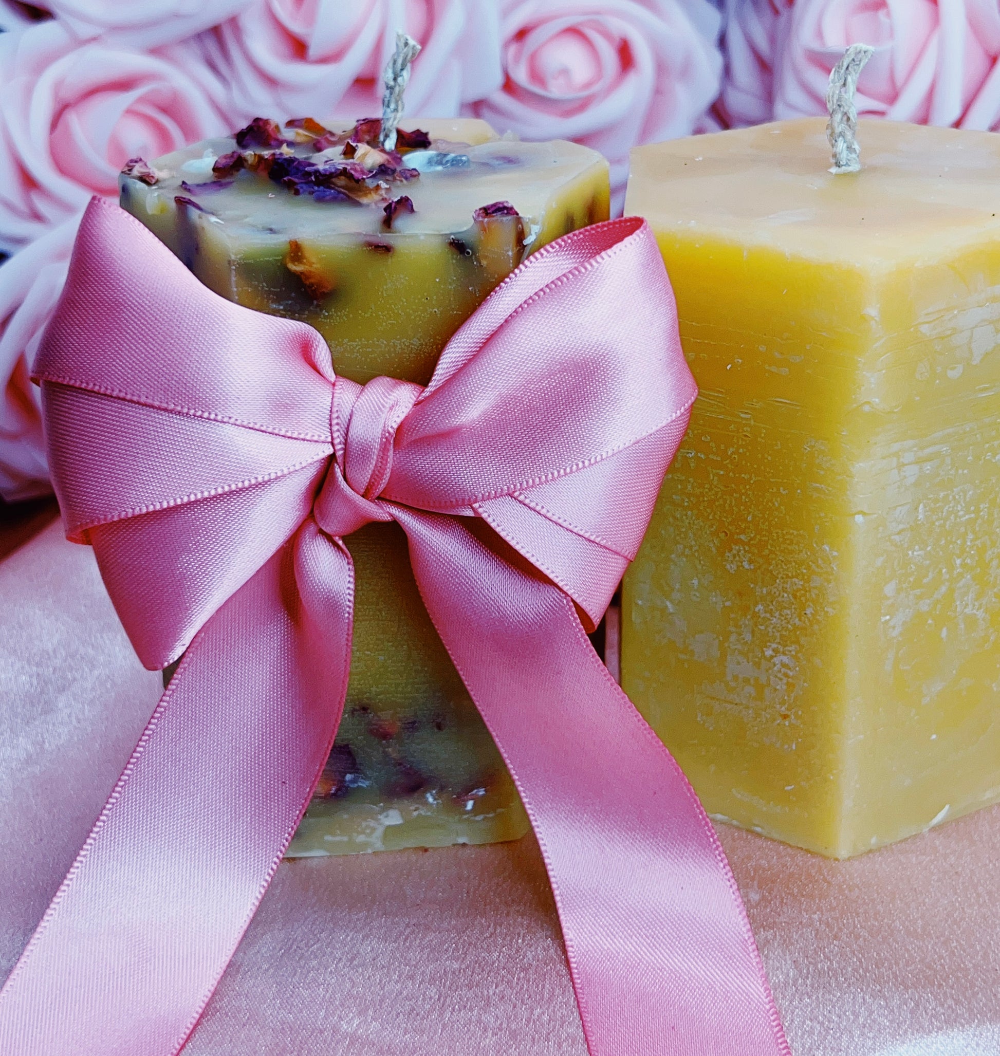 Two square candles with a pink ribbon and pink roses in the background