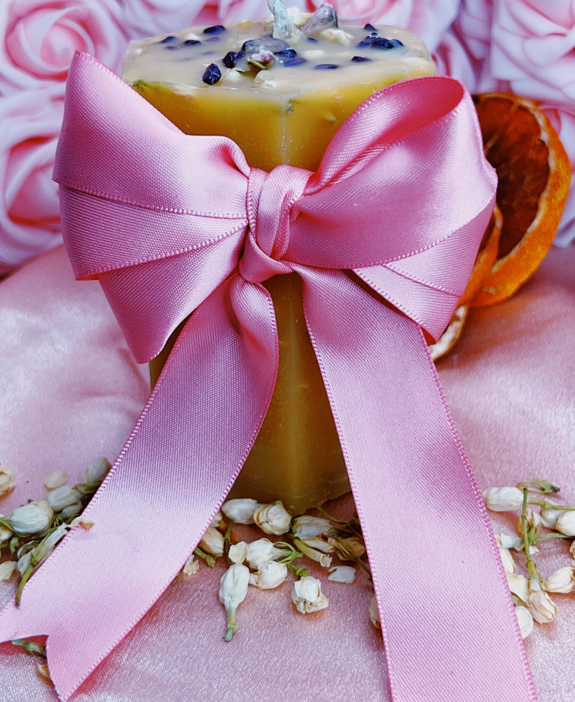 Yellow candle with a pink ribbon on a pink surface with flowers and an orange slice.