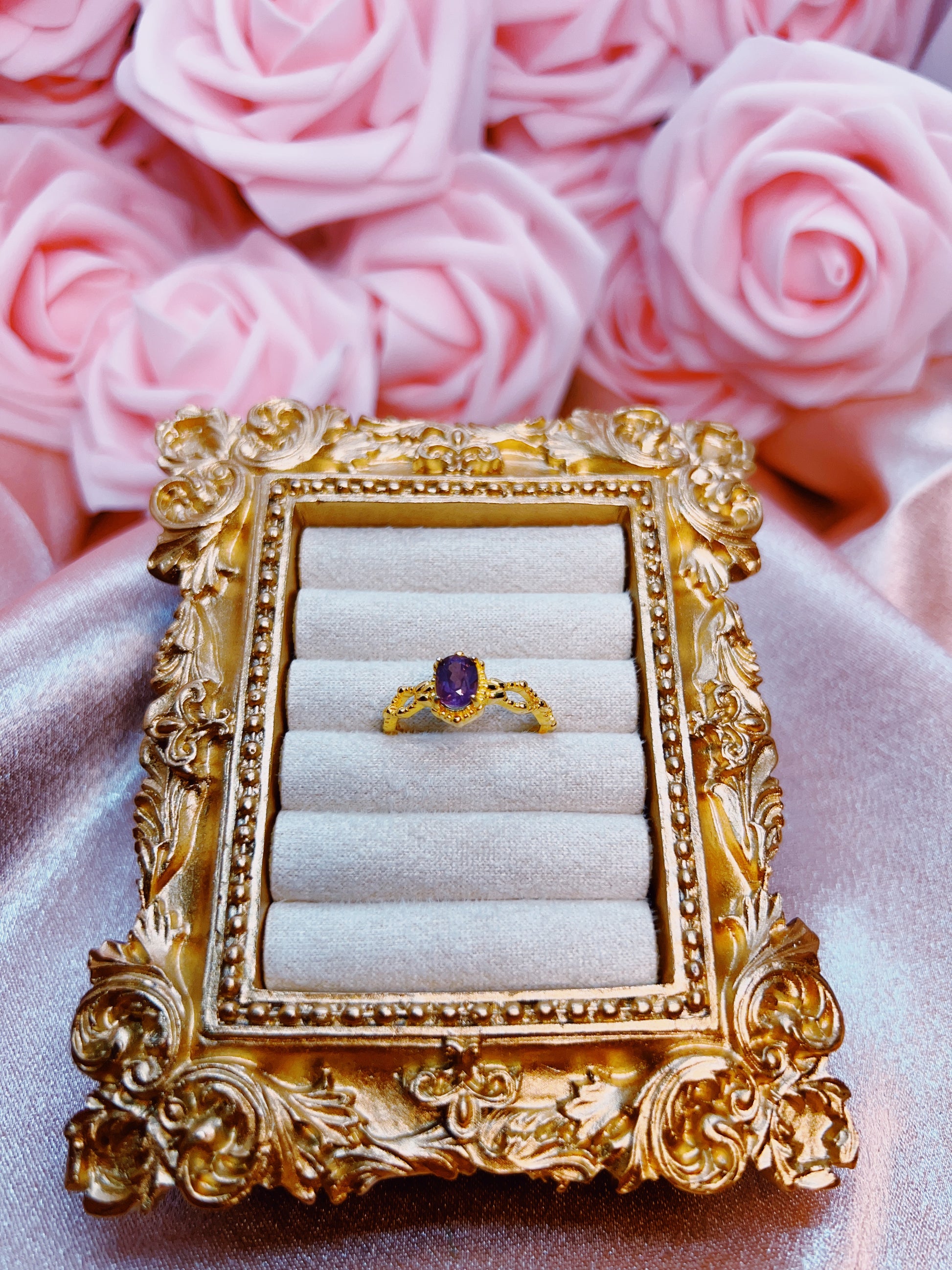 Gold ornate frame with a ring featuring a purple gemstone against pink roses.